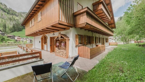 A two-story wooden house with a balcony, a stone wall, and a wooden deck with chairs. - I Folli di Alleghe Apartment - Parking (Caprile)