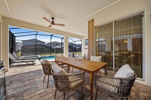 An outdoor patio with a wooden table and wicker chairs, a ceiling fan, and a view of a swimming pool and houses. - 6BR house sleeps 16 with private pool (Kissimmee)
