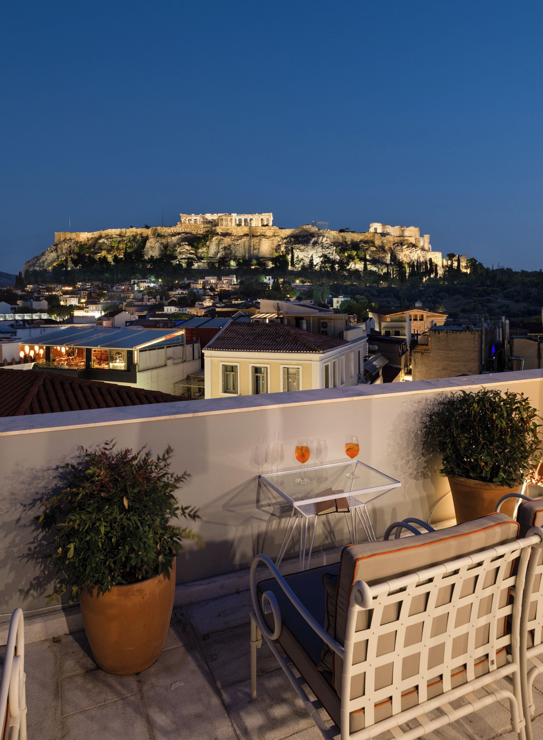 Eine Terrasse mit Tisch und Stühlen, mit Blick auf eine historische Stadtlandschaft und antike Ruinen auf einem Hügel.