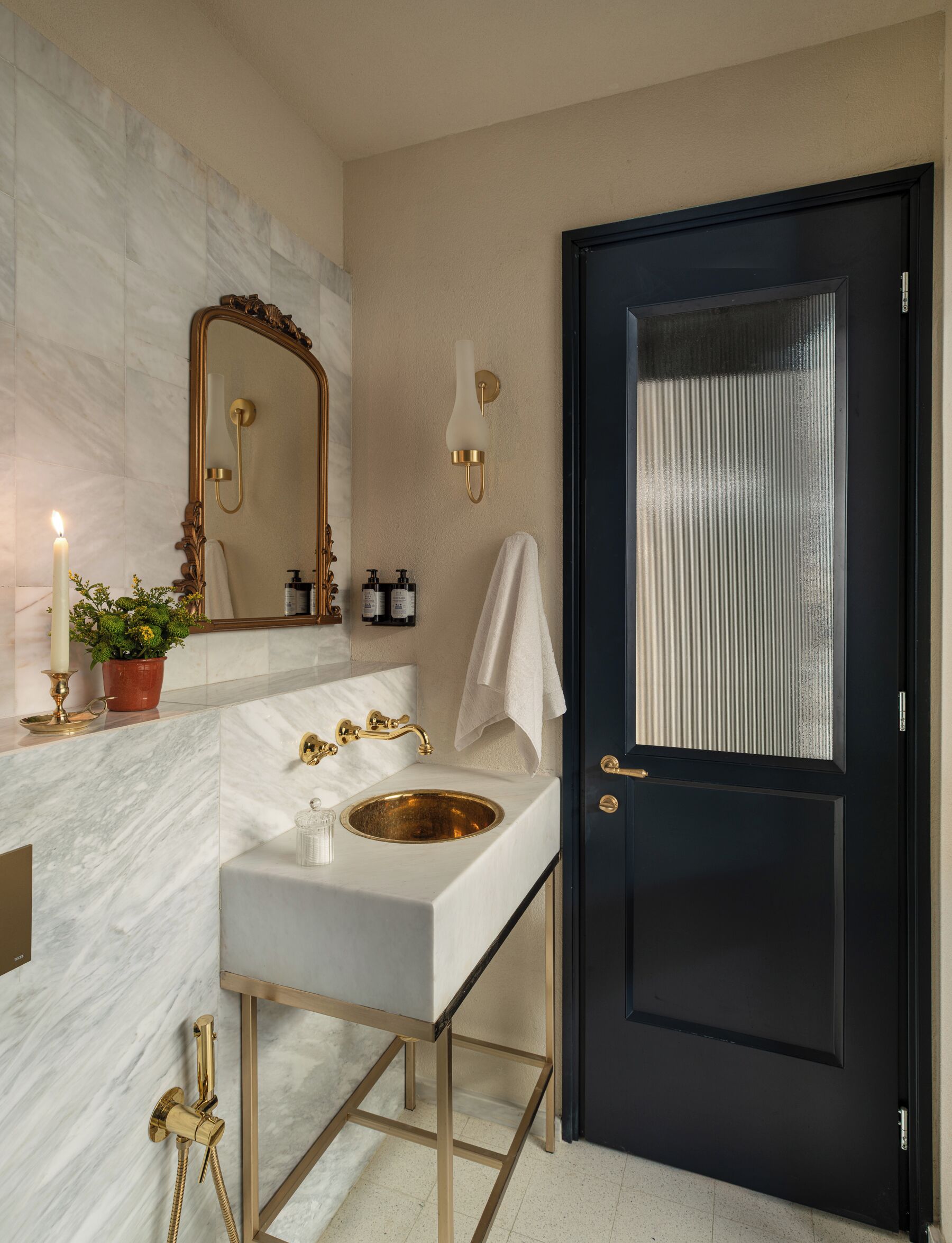 Une salle de bain avec un meuble-lavabo en marbre, des robinetteries dorées, un grand miroir, une porte noire et une plante en pot.