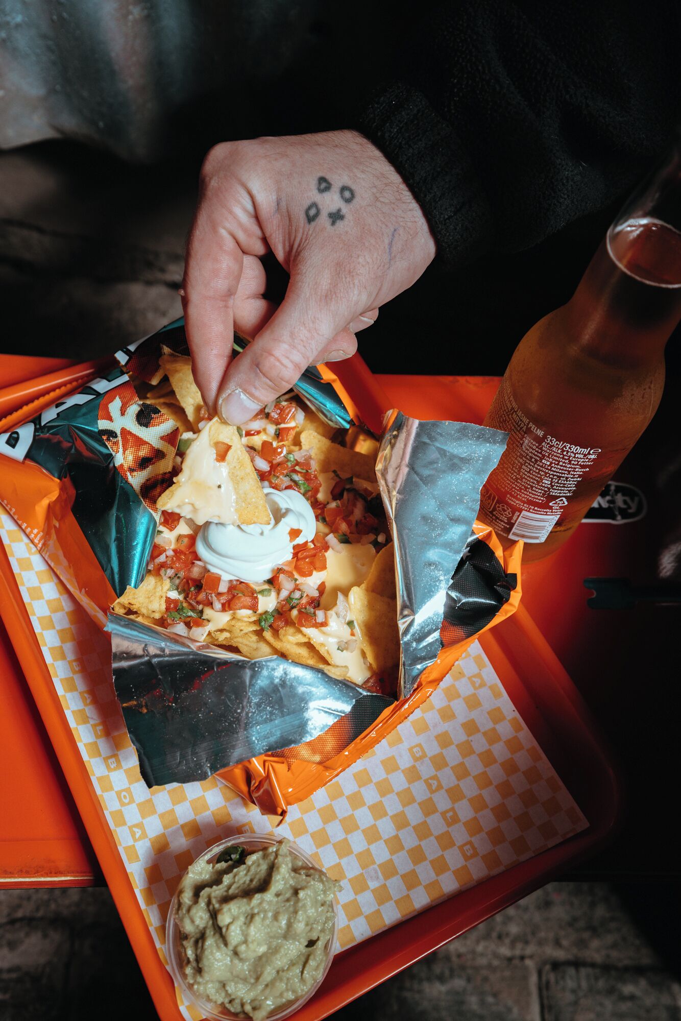 A hand holding a nacho dish with toppings, a drink bottle, and a side of guacamole on a tray.