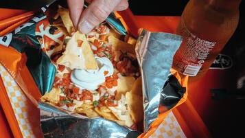 A hand holding a nacho dish with toppings, a drink bottle, and a side of guacamole on a tray.