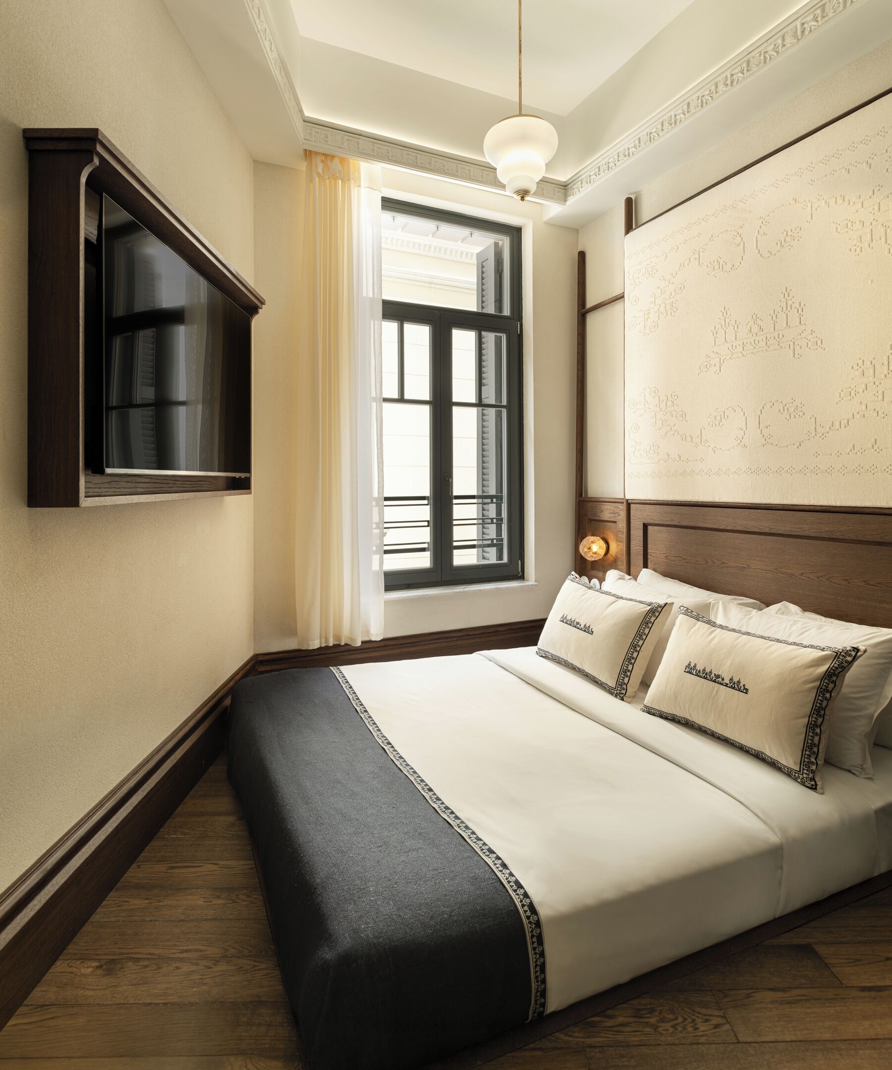 A hotel room with a bed, a TV mounted on the wall, a window with curtains, and a decorative headboard.