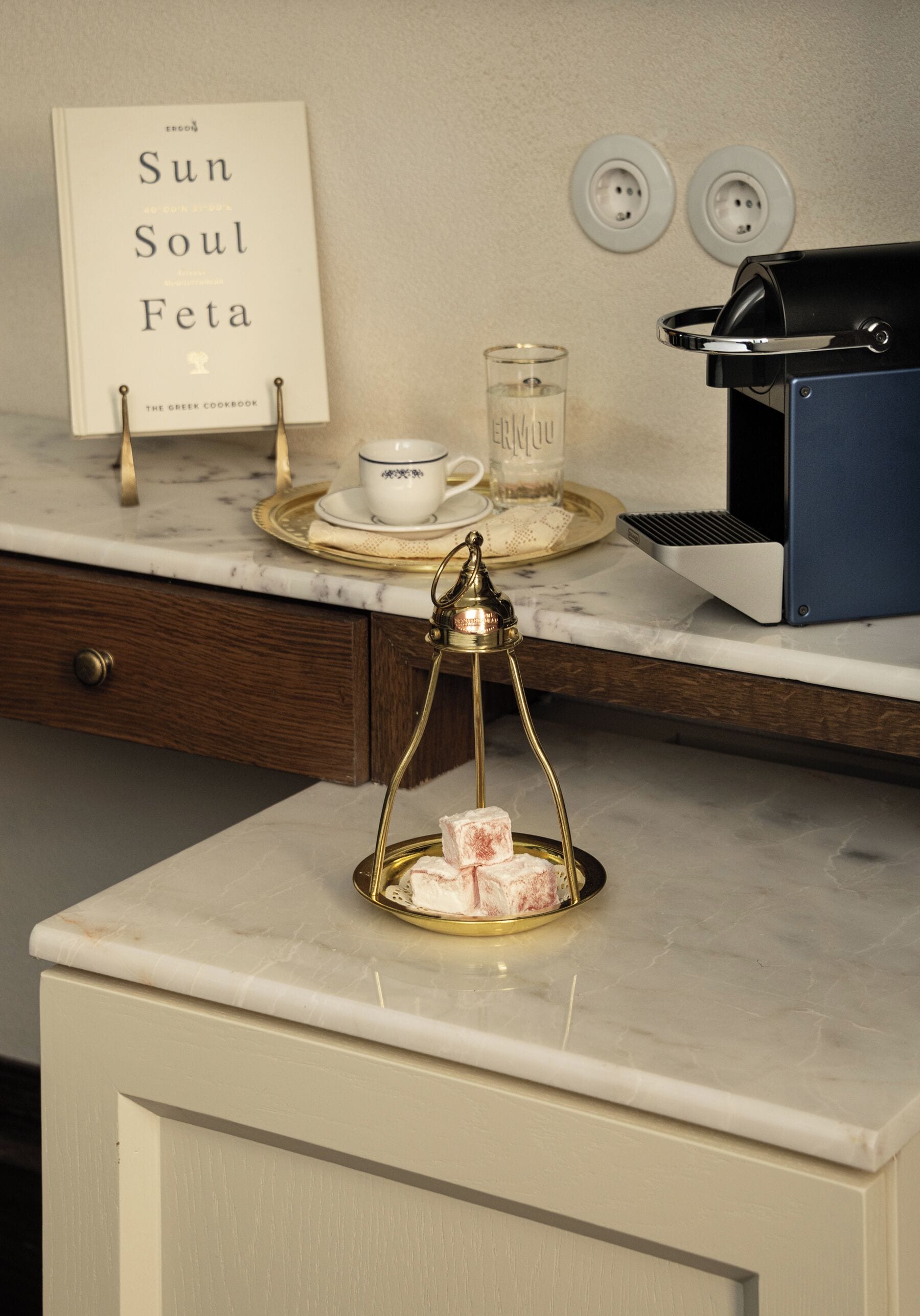 A kitchen counter with a book titled 'Sun Soul Feta,' a coffee machine, a glass with a candle, and a tray with food items.