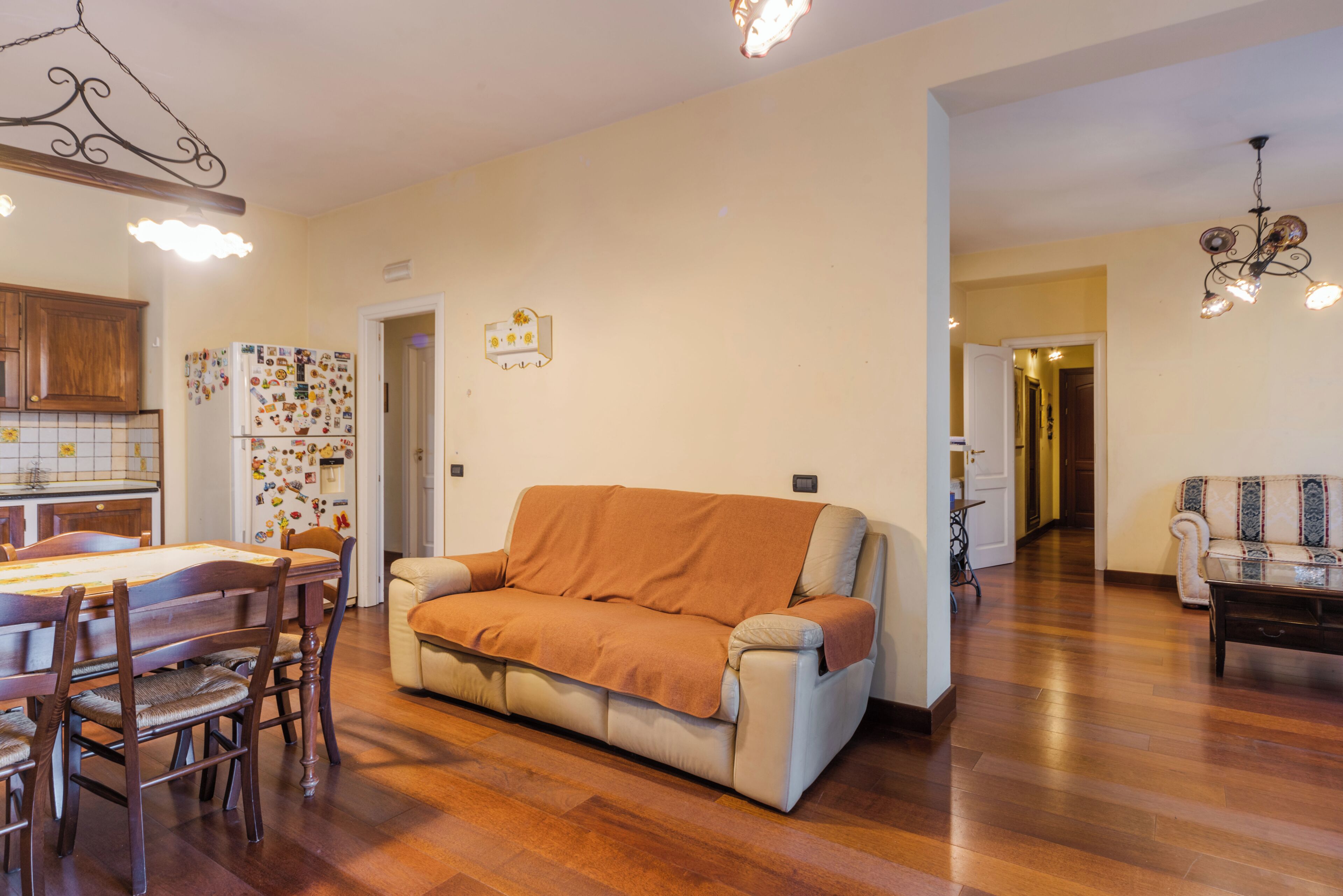 A living room with a sofa, a dining table with chairs, a kitchen area with cabinets and a refrigerator, and a hallway leading to another room.
