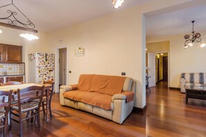 Een woonkamer met een bank, een eettafel met stoelen, een keuken met kasten en een koelkast, en een gang die naar een andere kamer leidt.