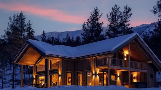 Una cabaña de madera grande de dos pisos, con amplias ventanas y un balcón, rodeada de nieve y árboles.