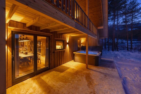 A wooden cabin with a glass door entrance, a hot tub, and a dining area.