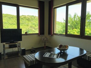 A room with a television on a stand, a round table with a placemat, a striped bench, and large windows with a view of greenery.