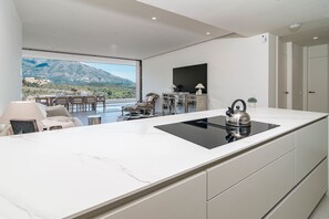 A modern kitchen with a large island, a dining area with chairs, a TV mounted on the wall, and a view of mountains. - RLQ - Modern 2 Bed Apt With Rooftop Plunge Pool (Benahavis)