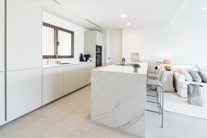 A modern kitchen with white cabinetry, a marble island, and a dining area with a sofa and a lamp. - RLQ - Modern 2 Bed Apt With Rooftop Plunge Pool (Benahavis)