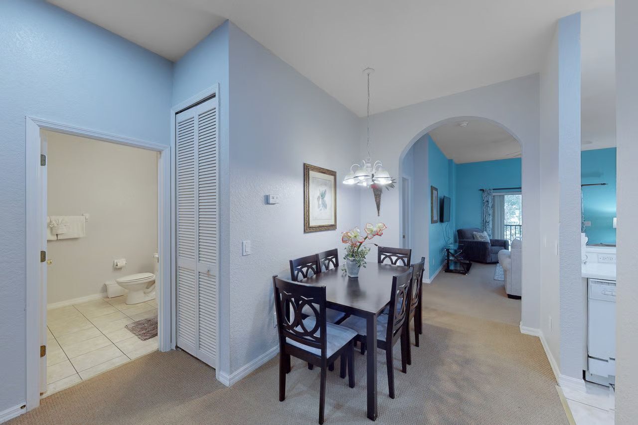 A dining area with a table and chairs, a hanging light fixture, a framed picture on the wall, and a view into the kitchen.
