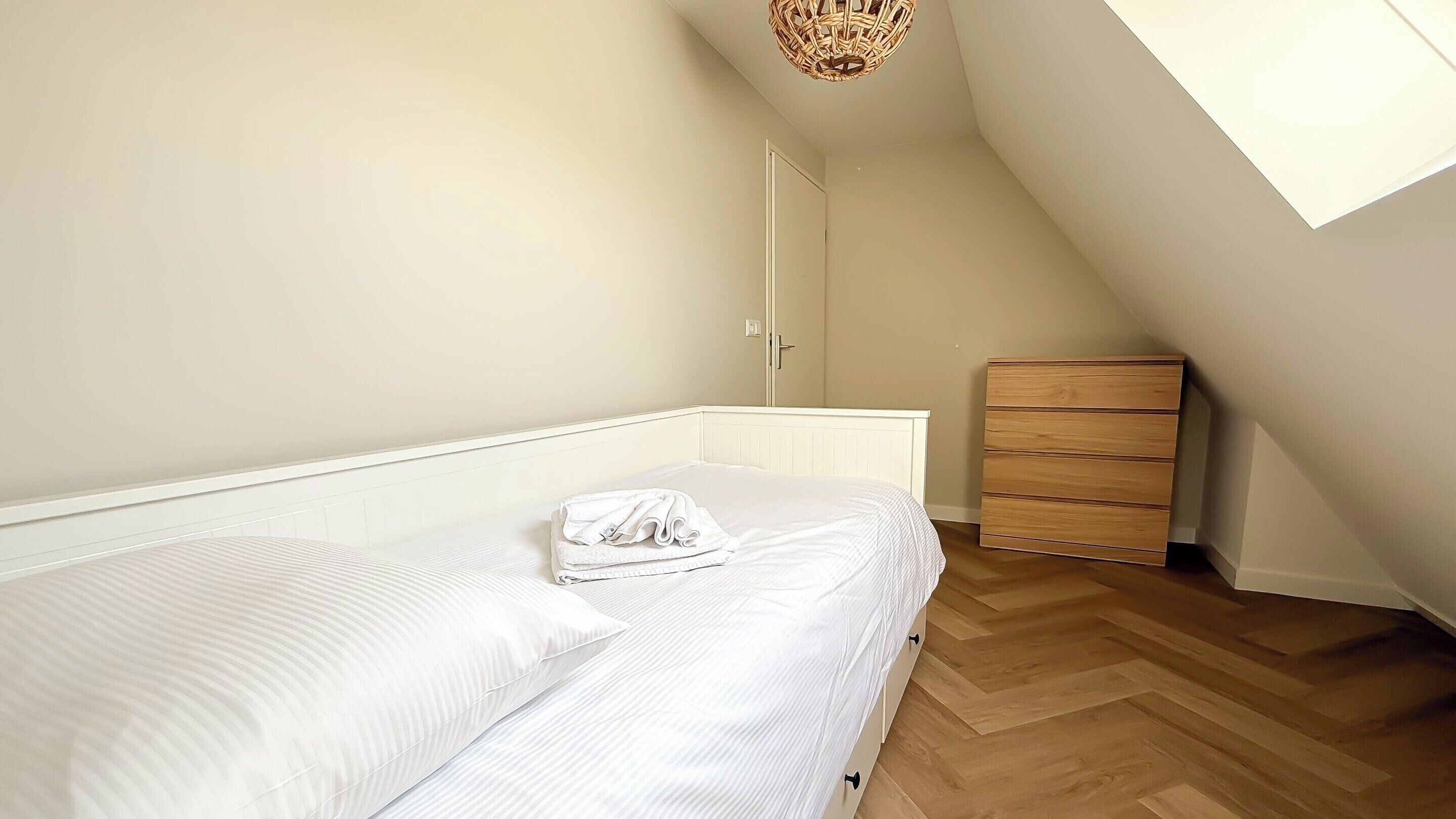 Una camera da letto con un letto, una cassettiera in legno, una porta e un soffitto spiovente.