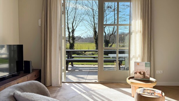 A room with a TV, a wooden coffee table, and a view of an outdoor area with a bench and picnic table. - Villa With Expansive Sunny Garden (Egmond aan den Hoef)