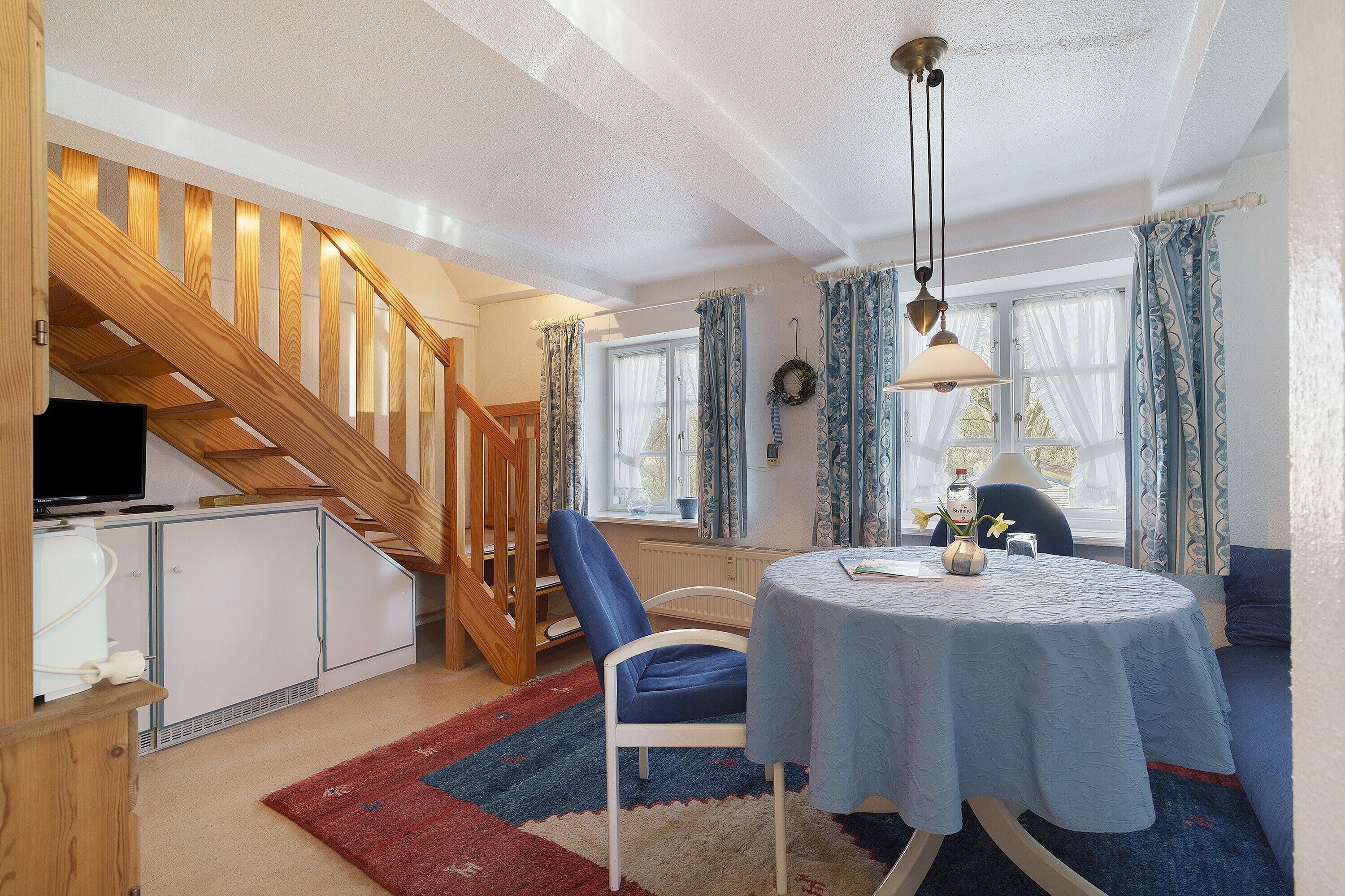 Ein Zimmer mit einer Holztreppe, einem Esstisch mit blauer Tischdecke, einem blauen Stuhl und einem Fernseher auf einem Schrank.