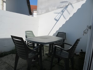Eine Terrasse mit Tisch und vier Stühlen, eine weiße Wand und der Schatten eines Gebäudes.