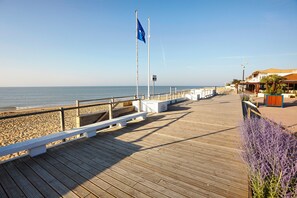 A wooden boardwalk leading to a beach with a flag, benches, and a building with a red roof. - Maisonnette de Martin, 50 m from the Beach (La Tranche-sur-Mer)