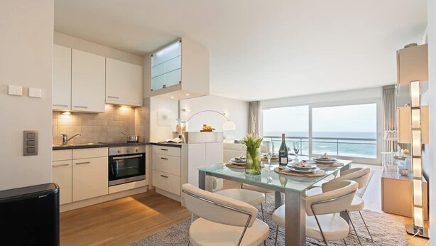Eine moderne Küche mit weißen Schränken, ein Essbereich mit einem Glastisch und Blick aufs Meer.