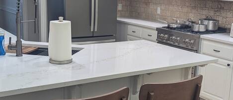 Una cocina moderna con armarios blancos, electrodomésticos de acero inoxidable, una isla central con dos taburetes de bar y un salpicadero de azulejos.