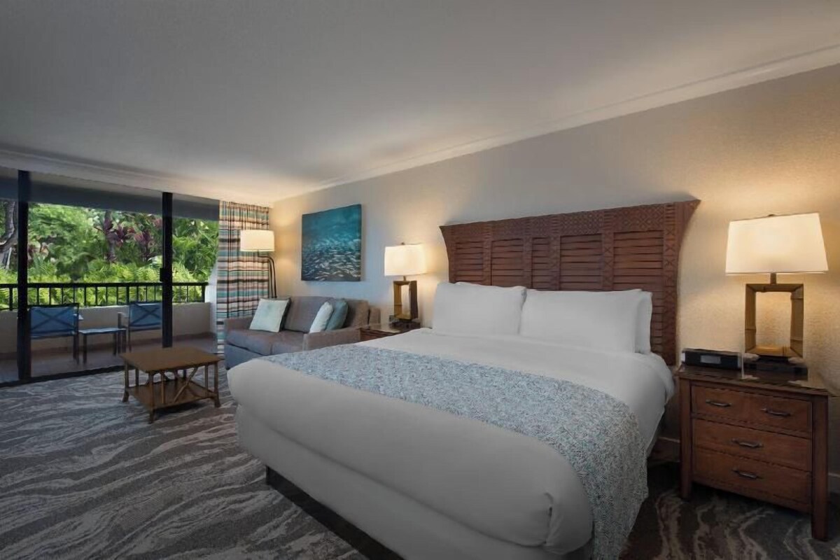 A hotel room with a large bed, two bedside tables with lamps, a sofa, a small table, and a view of greenery through large windows.