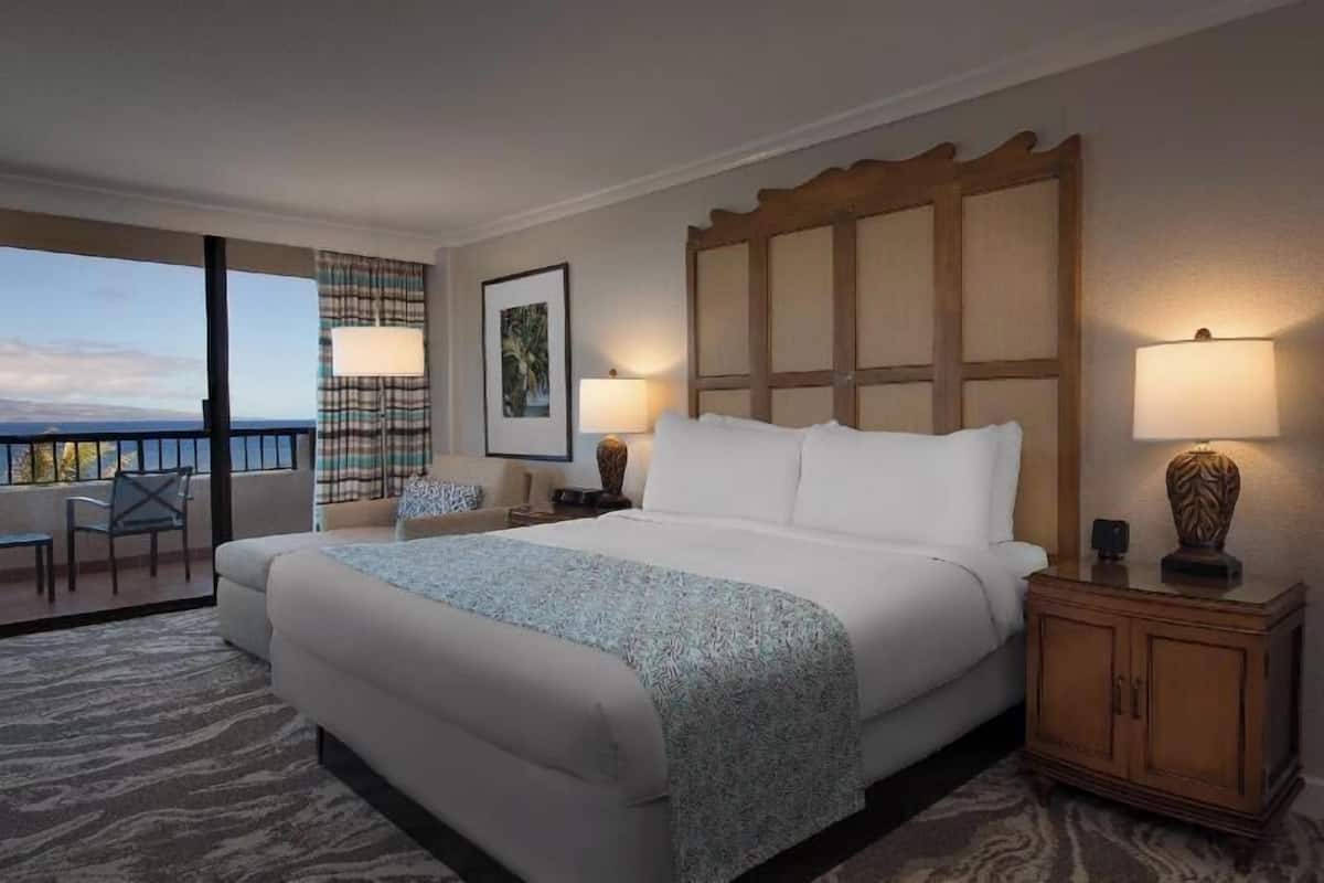 Ein Hotelzimmer mit einem großen Bett, einem Nachttisch mit Lampe, einem Sessel, einem Bild an der Wand und Blick aufs Meer.
