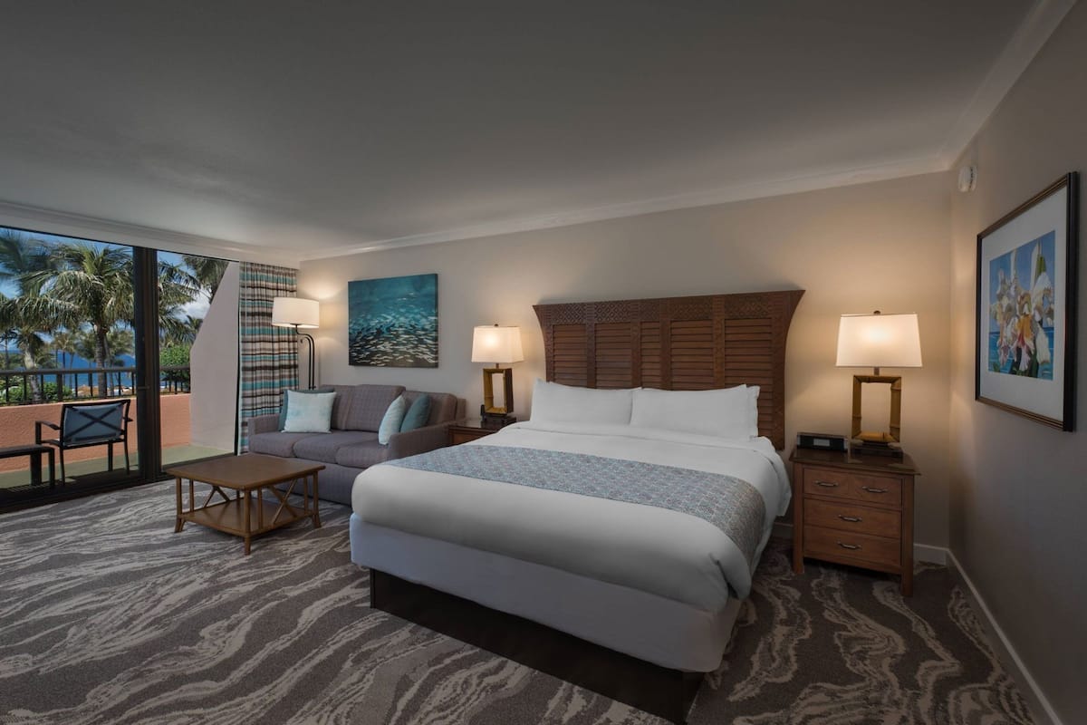 Ein Hotelzimmer mit einem großen Bett, einem Schlafsofa, einem kleinen Tisch, einem Nachttisch und einem Balkon mit Blick auf Palmen.