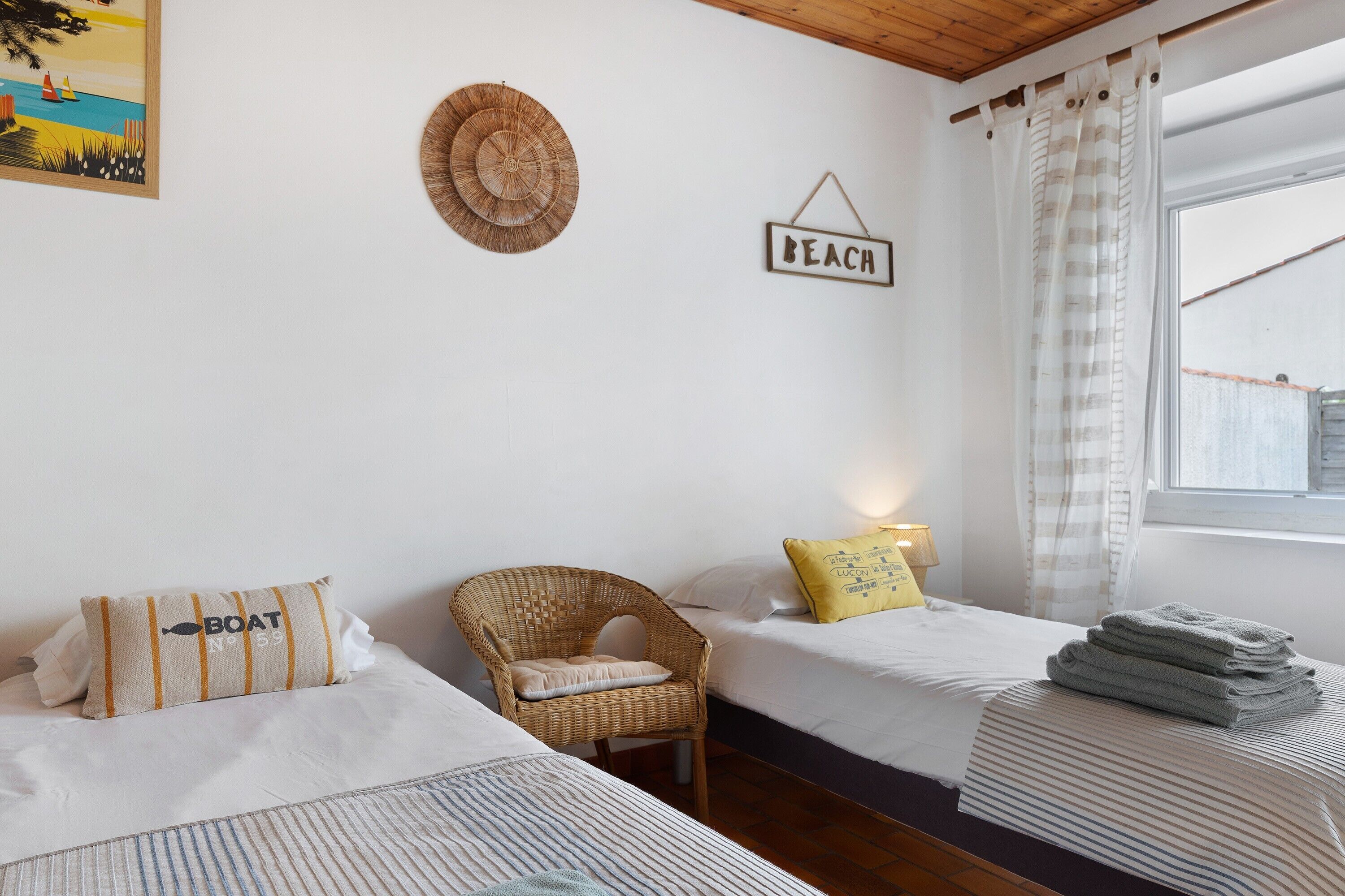 A bedroom with two beds, a wicker chair, a 'BEACH' sign, and a window with curtains.