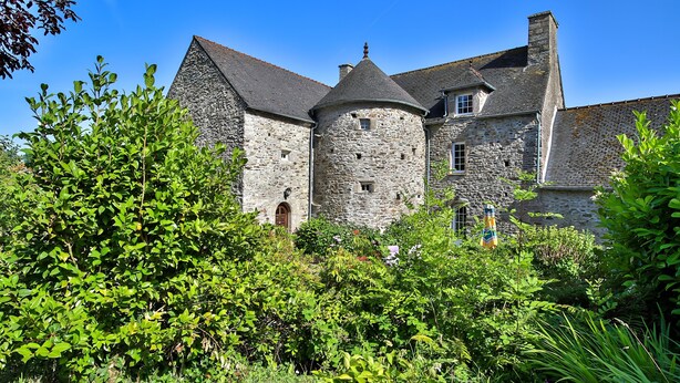 Un edificio de piedra con torrecilla y ventanas de arco, rodeado de abundante vegetación.