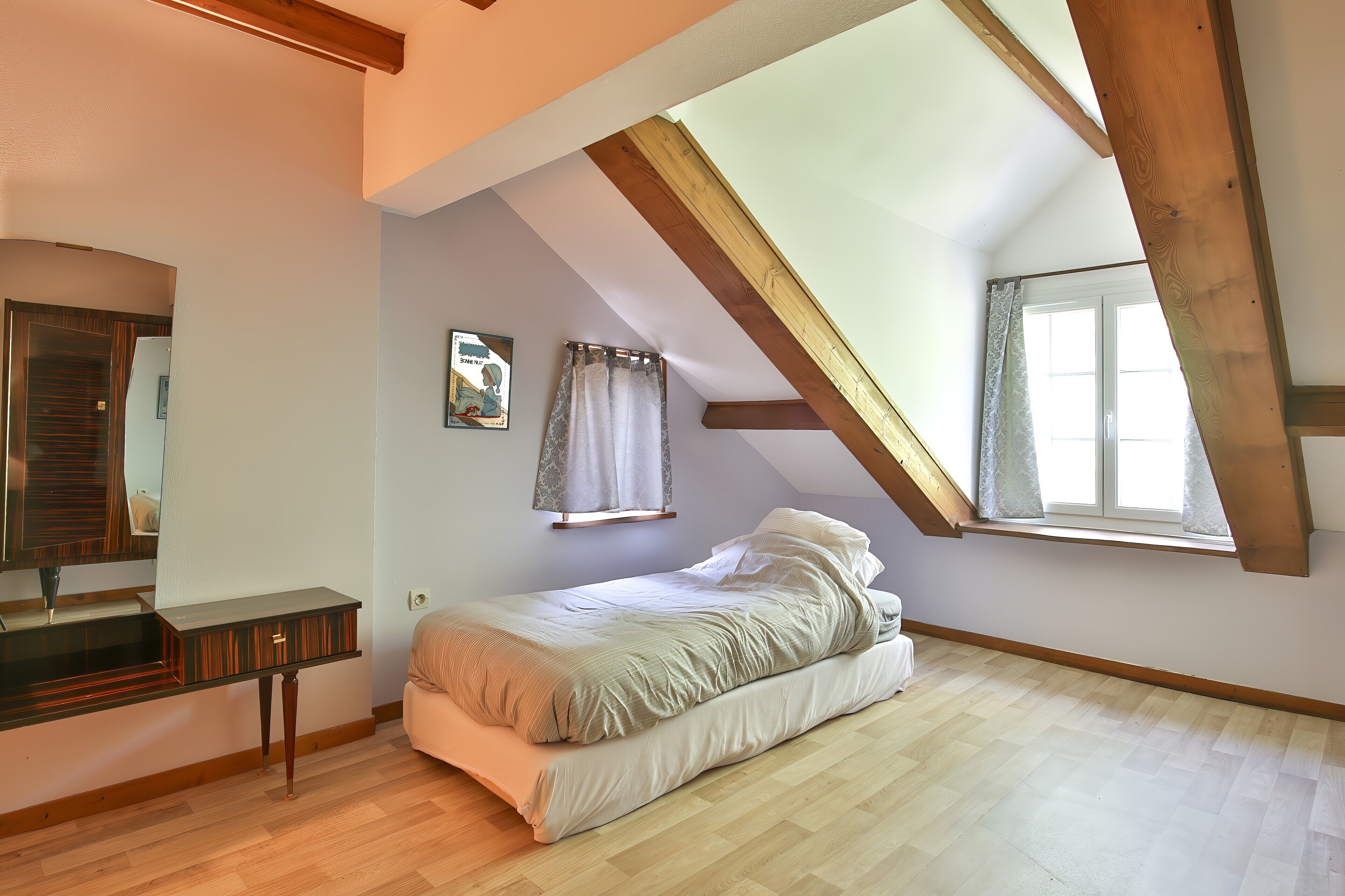 A bedroom with a sloped ceiling, a bed, a dresser, and a window with curtains.