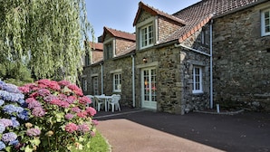 Un bâtiment en pierre avec un toit en tuiles, une allée pavée et un jardin agrémenté d’hortensias.