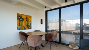 A modern room with a round wooden table, four upholstered chairs, a large window with a view of buildings and water, and a painting on the wall. - Stylish Boathouse on Lake Sneeker (Offingawier)