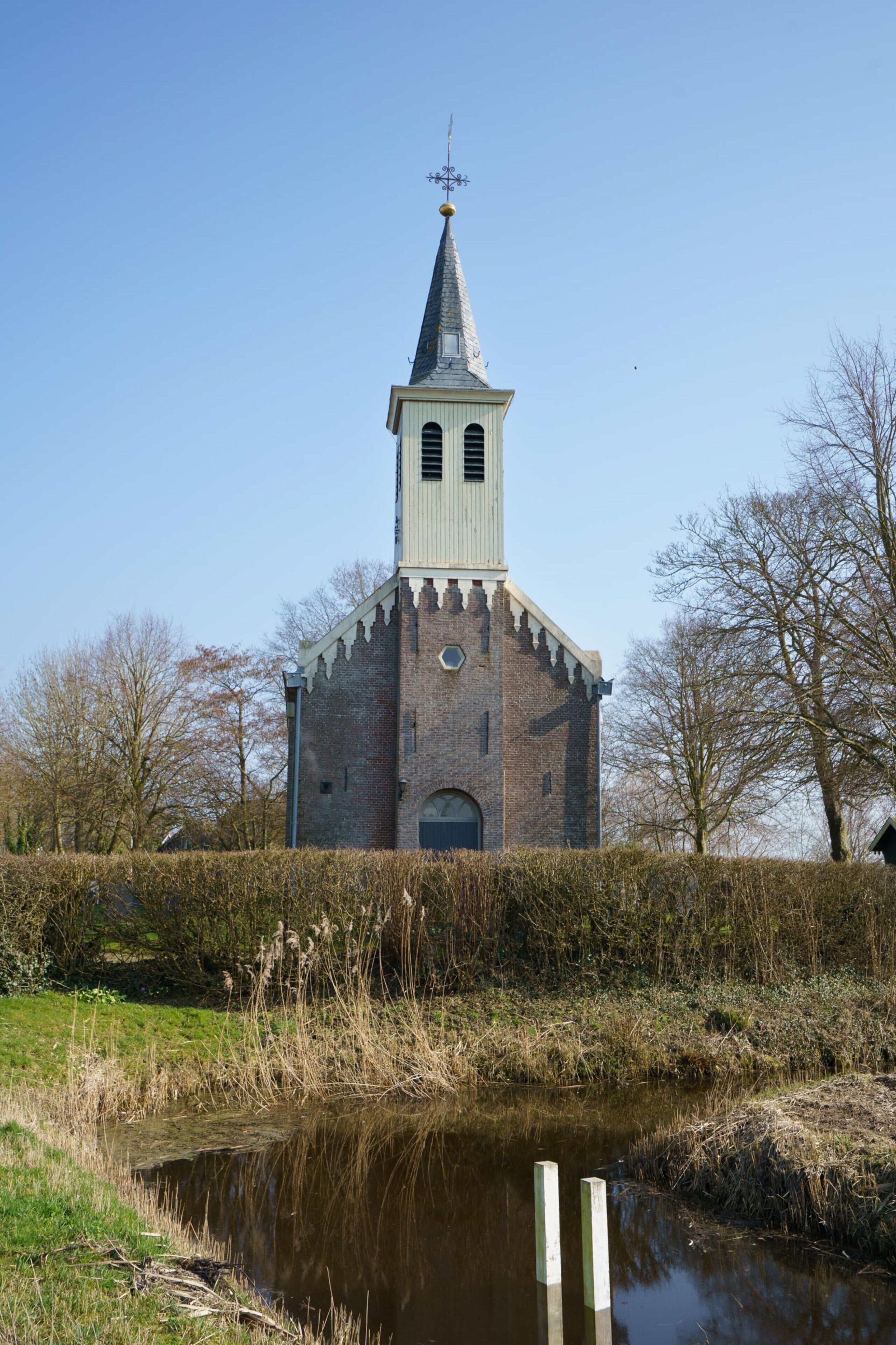 Eine Kirche mit einem hohen Turm, einem kleinen Teich davor und kahlen Bäumen im Hintergrund.