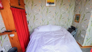 A single bed with a white duvet, a red curtain, a wooden shelf with a remote control, a small bucket, and a wall with floral wallpaper.