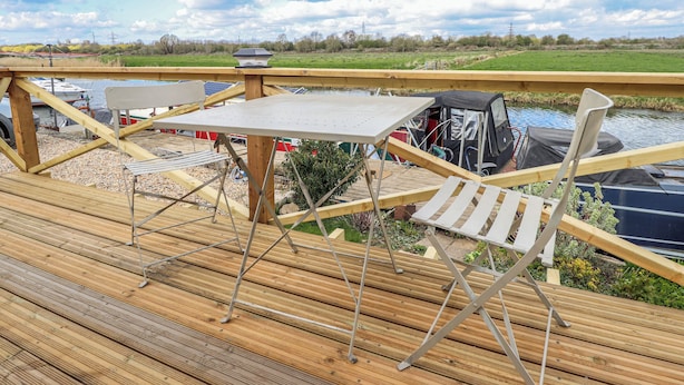 A wooden deck with a table and chairs, overlooking a body of water with boats and a distant landscape.