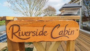 Uma placa de madeira com a inscrição 'Riverside Cabin' colocada sobre uma mesa de madeira ao ar livre.