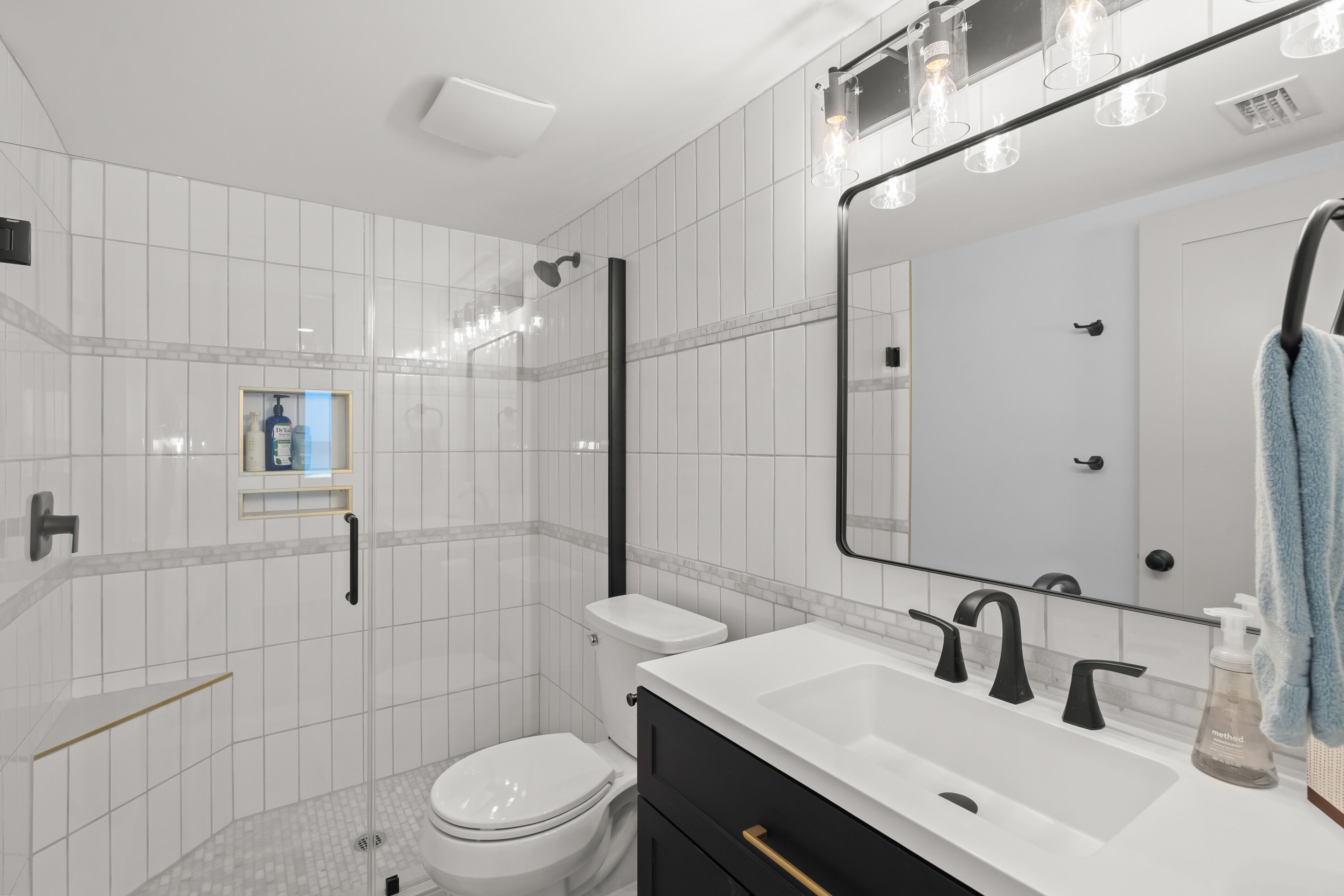 A modern bathroom with a white tiled wall, a glass shower enclosure, a white toilet, a black vanity with a mirror, and a towel hanging on the right.
