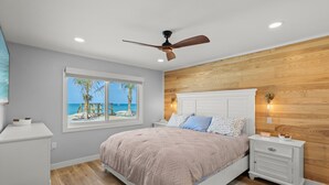 A bedroom with a bed, nightstands, a ceiling fan, a window with a view of palm trees, and a wooden wall panel. - Fully Remodeled First-Floor Condo | Steps to Beach | Siesta Key (Sarasota)