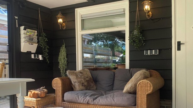A wicker sofa with cushions, a small round ottoman, and hanging plants against a dark wall with a window. - Quiet Wellness Villa in Bellingwolde (Bellingwolde)