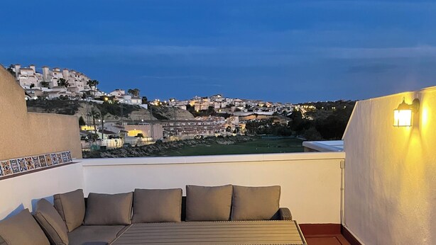 Eine Dachterrasse mit einer Couch, einem Tisch und Blick auf eine städtische Silhouette im Abendlicht.