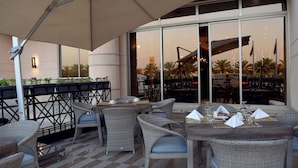 An outdoor seating area with wicker furniture and a checkered floor, overlooking a building with large windows and a view of palm trees. - Best Plus Aurora Hotel (Riyadh)