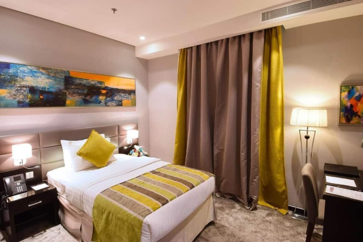 Una habitación de hotel con cama, mesitas de noche, escritorio, lámpara y un cuadro en la pared.
