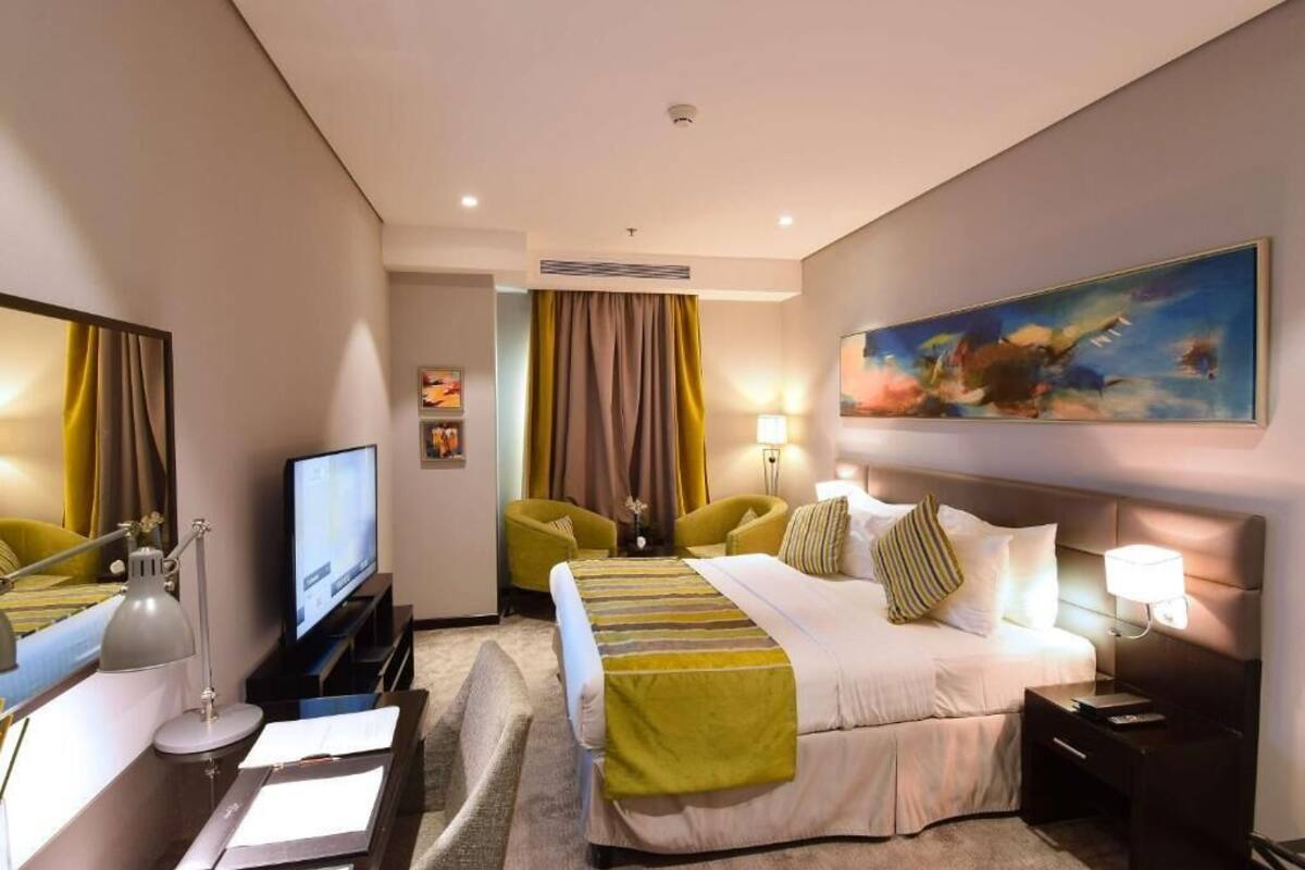 Una habitación de hotel con una cama, un escritorio con computadora, televisión, una silla y un cuadro en la pared.