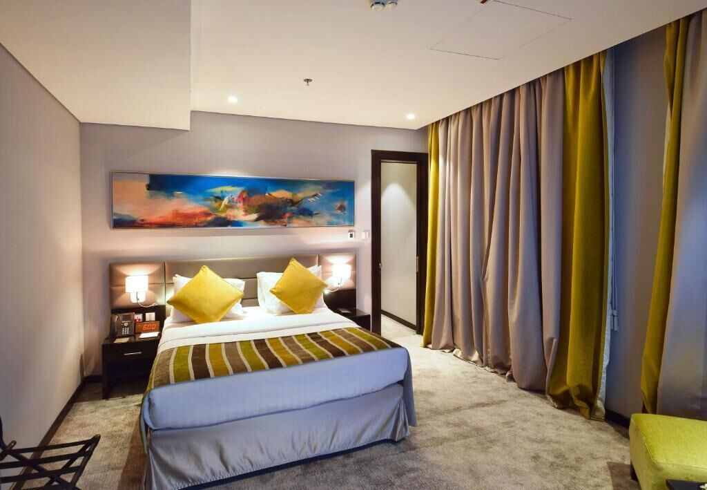 A hotel room with a bed, a bedside table with a lamp, a painting on the wall, and curtains.