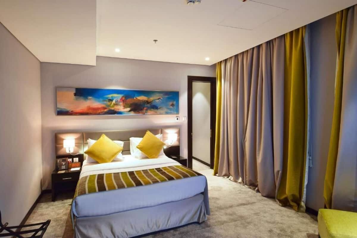 Una habitación de hotel con una cama, una mesita de noche con lámpara, un cuadro en la pared y cortinas.