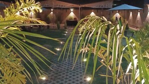 Zona esterna illuminata di notte con pavimento decorato, palme e strutture illuminate.