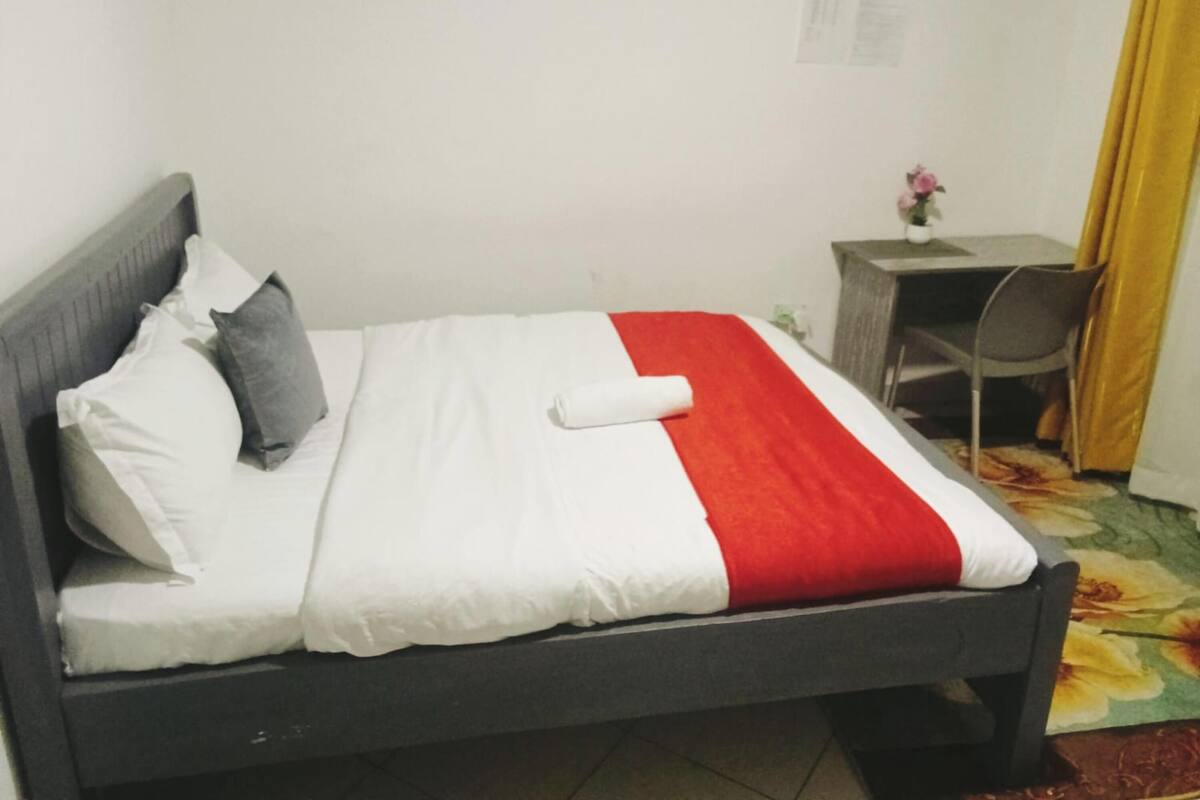 A single bed with a headboard, a desk with a chair, and a small table with a vase of flowers.