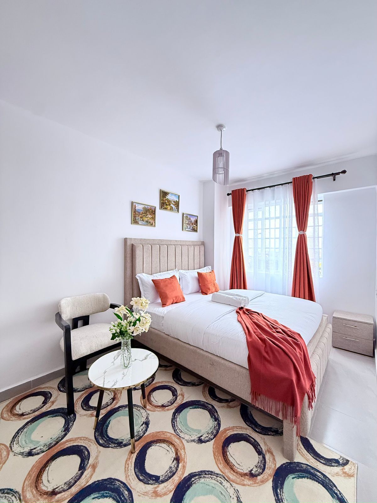 A bedroom with a bed, a chair, a small table with flowers, and a patterned carpet.