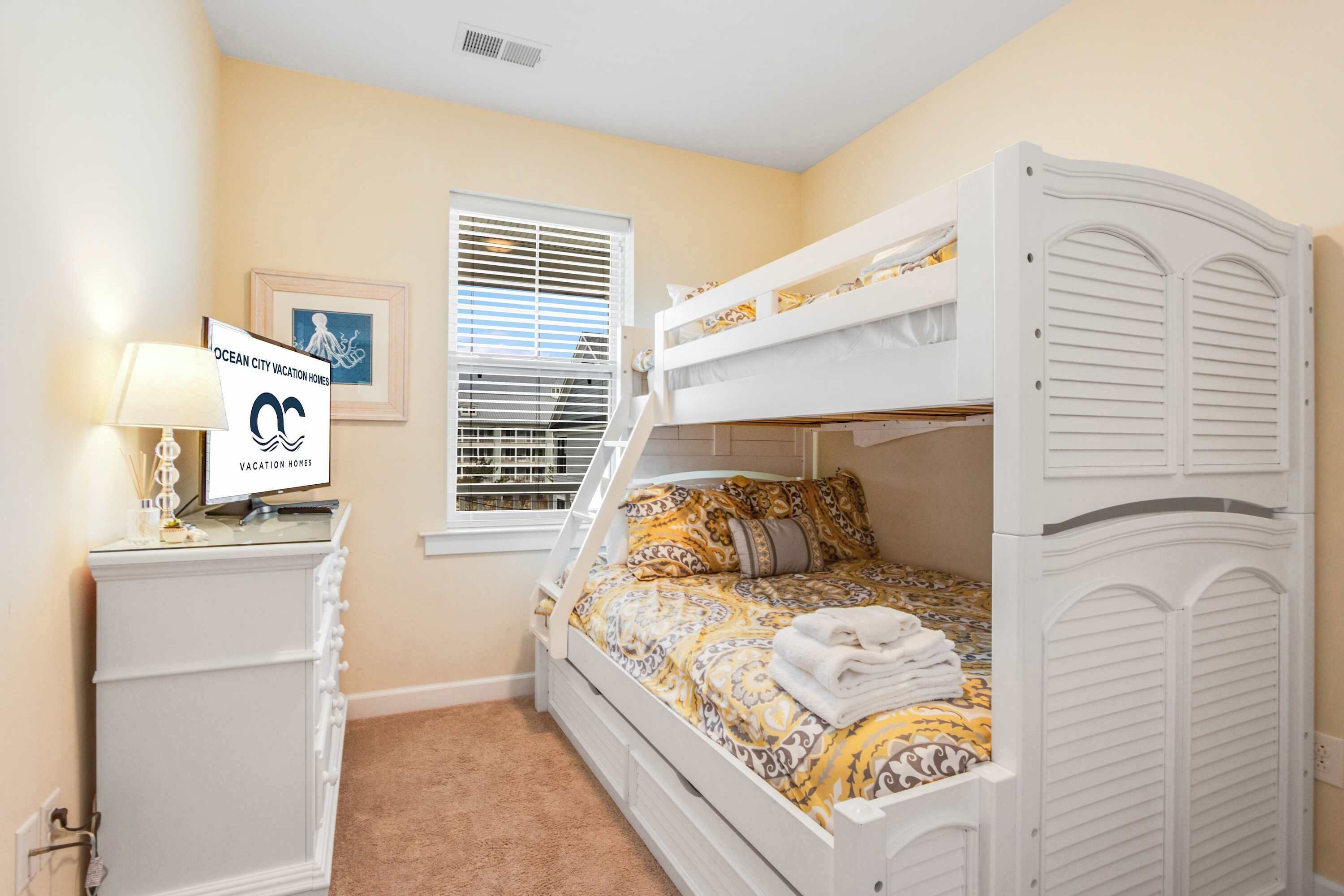 A bedroom with bunk beds, a bedside table with a lamp, a framed picture, and a window with blinds.