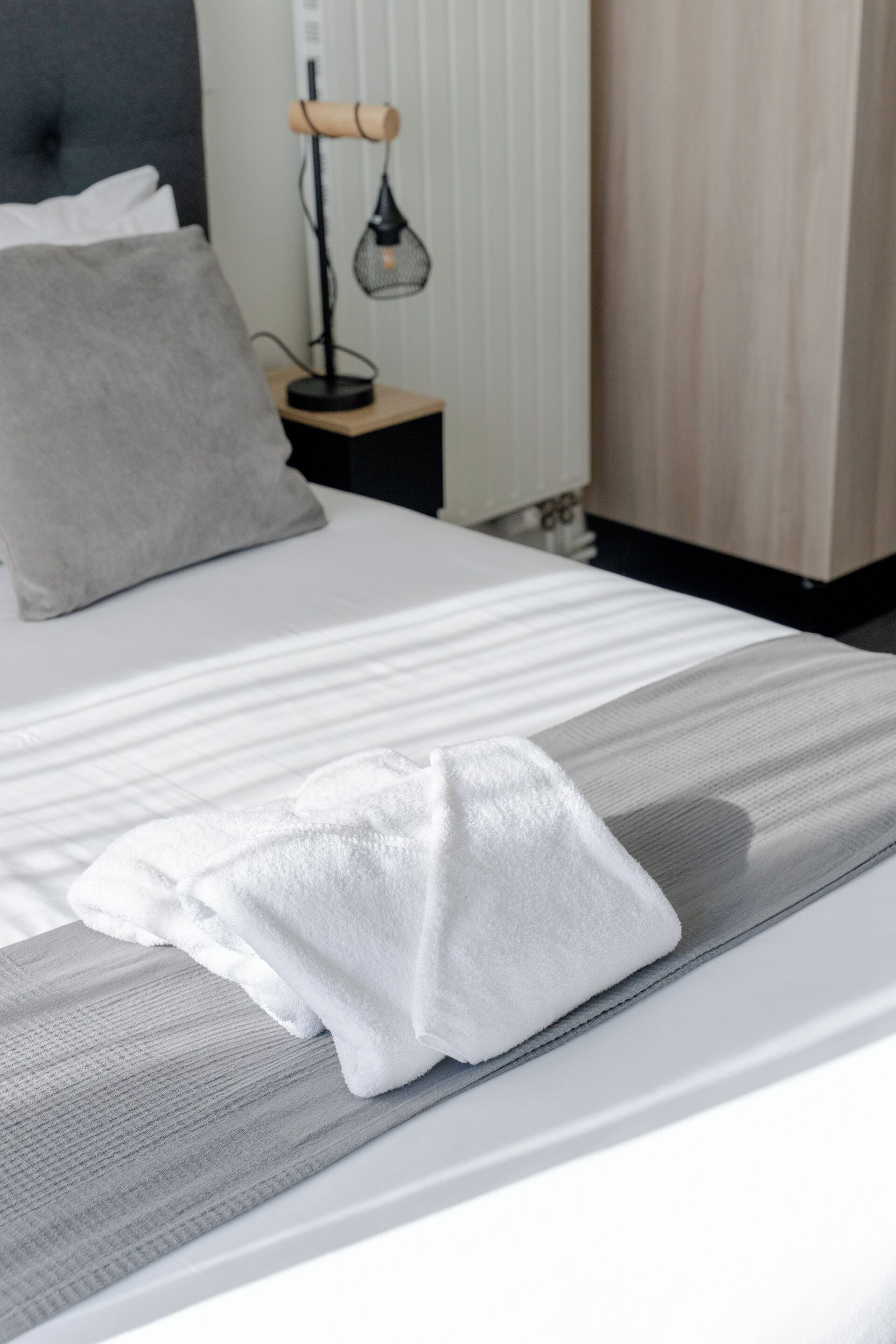 Ein ordentlich bezogenes Bett mit einer grau-weißen Streifenauflage, einem weißen Kopfkissen und einem weißen Handtuch.