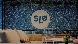 Un área de descanso moderna con un sofá, una mesa redonda y una silla. En la pared, se observa un letrero circular con las letras 'SLO'.